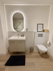 Ein modernes Badezimmer mit einem Waschbecken und einem großen, beleuchteten Spiegel. Die Wände sind hell und der Boden ist aus Holz.