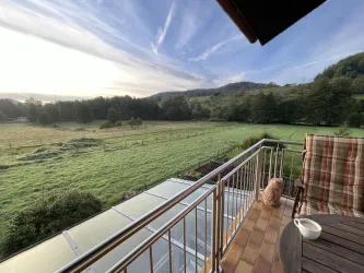 Eine Aussicht von einem Balkon auf eine grüne Wiese und sanfte Hügel. Im Vordergrund stehen ein Stuhl und eine Tasse.