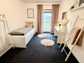 Ein modernes Schlafzimmer mit einem Einzelbett, rosa Vorhängen und einem großen Fenster. Es gibt eine gemütliche Dekoration mit zwei runden Teppichen und einem Spiegel.