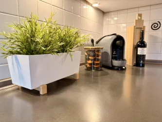 Eine moderne Küchenecke mit einer Pflanze in einem weißen Topf, einer Kaffeemaschine und einer Glasbox mit Keksen. Im Hintergrund steht eine Flasche Wein und ein Schneidebrett.