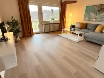 Helles, modern eingerichtetes Wohnzimmer mit großer Fensterfront und gemütlicher Sitzlandschaft. Warme Beleuchtung, stilvolle Dekoration und ein schöner Ausblick ins Grüne schaffen eine entspannte Wohlfühlatmosphäre