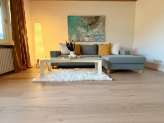 Ein modernes Wohnzimmer mit einem grauen Sofa und einem eleganten Couchtisch. Helle Farben und dekorative Elemente schaffen eine gemütliche Atmosphäre.