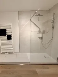 Eine moderne Dusche mit Glastüren in einem hellen Badezimmer. An der Wand hängen Handtücher und der Boden ist aus Holz.