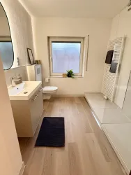 Ein modernes Badezimmer mit hellem Holzfußboden und einem großen Fenster. Die Ausstattung umfasst ein Waschbecken, eine Toilette und eine Dusche.