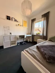 Ein modernes Schlafzimmer mit einem Einzelbett und einem Schreibtisch. Große Fenster lassen viel Tageslicht herein und die Wände sind in hellen Farben gehalten.