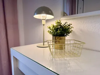 Ein moderner Tisch mit einer Lampe und einer kleinen Pflanze in einem goldenen Topf. Daneben steht ein goldener Korb auf der glatten Oberfläche.