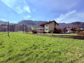 Ein großes, grünes Feld mit einem charmanten Haus im Hintergrund. Die Landschaft ist von sanften Hügeln und einem klaren blauen Himmel umgeben.