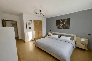 Ein modernes Schlafzimmer mit einem großen Bett und eleganter Bettwäsche. Die Wände sind grau, und es gibt Kunstwerke sowie Holzdetails, die eine gemütliche Atmosphäre schaffen.