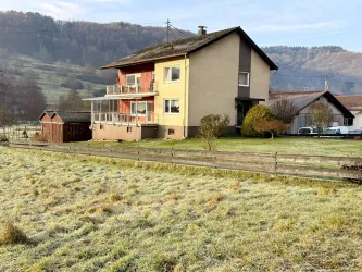 Ein zweigeschossiges Wohnhaus mit einem Balkon steht in einer ländlichen Umgebung. Der Garten ist grün und von einer Zaun umgeben.