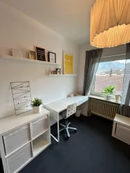 Ein modernes Arbeitszimmer mit einem Schreibtisch, einem Stuhl und Regalen an der Wand. Große Fenster sorgen für viel Tageslicht und eine angenehme Atmosphäre.
