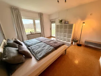 Ein helles Schlafzimmer mit einem großen Bett und großen Fenstern. Eine schlichte Regaleinheit und eine Stehlampe ergänzen die gemütliche Atmosphäre.