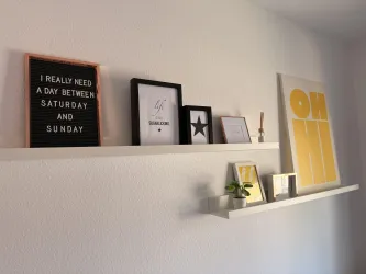 Eine schlichte Wand mit zwei Regalen, die mit verschiedenen Bilderrahmen und einer kleinen Pflanze dekoriert sind. Auf dem oberen Regal befindet sich ein Schriftzug, der den Wunsch nach einem zusätzlichen Tag zwischen Samstag und Sonntag ausdrückt.