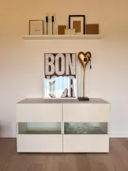 Ein moderner Wohnbereich mit einem weißen Sideboard und einer dekorativen Lampe. An der Wand hängt ein großes Bild mit dem Wort "BON" und einige gerahmte Bilder.
