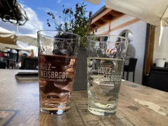 Zwei Gläser stehen auf einem Holztisch, eines mit rosafarbenem Getränk und das andere mit klarem Wasser. Im Hintergrund sind Pflanzen und eine entspannte Außenatmosphäre sichtbar.