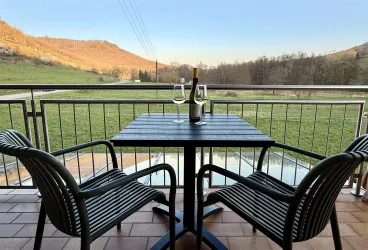 Ein Balkon mit zwei Stühlen und einem Tisch. Auf dem Tisch stehen eine Flasche Wein und zwei Gläser, mit Blick auf eine grüne Wiese.
