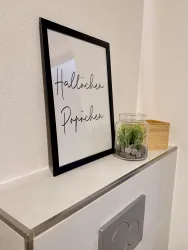 Ein Rahmen mit dem Aufdruck „Hallöchen Püppi“ steht auf einer Oberfläche. Daneben befindet sich ein Glas mit künstlichem Gras und ein kleiner Holzkasten.