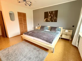 Ein modernes Schlafzimmer mit einem großen Bett und einem weichen Teppich. An der Wand hängt ein Kunstwerk, und durch das Fenster fällt natürliches Licht.