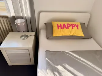 Ein einfaches Schlafzimmer mit einem Bett und einem Nachttisch. Auf dem Bett liegt ein leuchtend gelbes Kissen mit der Aufschrift "HAPPY".