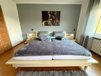 Ein modernes Schlafzimmer mit einem Doppelbett und dekorativen Kissen. An der Wand hängt ein Kunstwerk, und das Zimmer hat große Fenster mit Vorhängen.