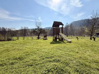 Ein Spielplatz mit Rutsche steht in einem grünen Garten. Im Hintergrund sind sanfte Hügel und ein blauer Himmel zu sehen.