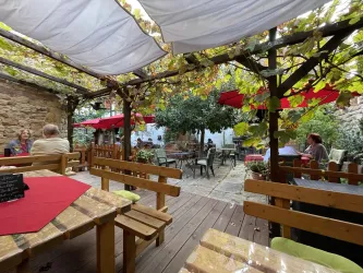 Ein gemütlicher Innenhof mit Holzmöbeln und roten Sonnenschirmen. Überdachte Plätze und grüne Pflanzen schaffen eine einladende Atmosphäre.