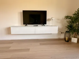 Ein moderner Wohnraum mit einem an der Wand montierten Fernseher und einem schlichten weißen TV-Schrank. Daneben steht eine Pflanze in einem Topf und eine Tischlampe.