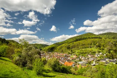 Eine malerische Landschaft mit einem kleinen Dorf inmitten von grünen Hügeln. Weißer Wolkenhimmel über einer ruhigen, idyllischen Umgebung.