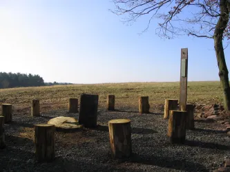 Ein rundes Holzlager mit Sitzstämmen steht auf einem offenen Feld. Im Hintergrund ist ein klarer Himmel und Bäume am Horizont sichtbar.