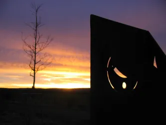 Ein schöner Sonnenuntergang mit leuchtenden Farben am Himmel. Im Vordergrund steht eine kunstvolle Skulptur und ein einzelner Baum.