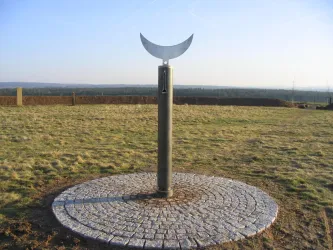 Ein metallisches Kunstwerk in Form einer Halbmondskulptur auf einem Steinfundament. Die umgebende Landschaft ist weitläufig und offen.