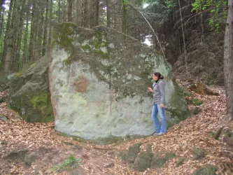 Eine Frau steht neben einem großen Felsen im Wald. Der Boden ist mit Laub bedeckt, und die Umgebung ist von Bäumen umgeben.