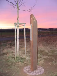 Eine metallische Skulptur steht auf einem runden Steinpflaster. Im Hintergrund ist ein schöner Sonnenuntergang mit lila und orangen Farben zu sehen.