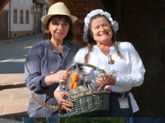Zwei Frauen stehen in einer malerischen Straße. Sie halten einen Korb mit handgefertigten Figuren und lachen freundlich.