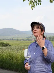 Eine Frau hält ein Weinglas und spricht mit gestikulierenden Händen. Im Hintergrund sind sanfte Hügel und eine grüne Landschaft zu sehen.