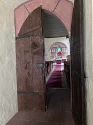 Eine offene Holztür führt in eine Kirche mit einer roten Läufer und einem schönen Fenster. Der Innenraum ist schlicht und einladend.