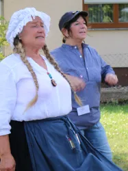 Zwei Frauen stehen nebeneinander und scheinen zu tanzen oder zu singen. Eine Frau trägt ein traditionelles Outfit mit Zöpfen, während die andere lässig gekleidet ist.