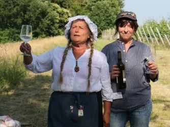 Zwei Frauen stehen in einem Weinberg und heben Gläser. Die eine trägt ein traditionelles Kostüm, während die andere lässige Kleidung anhat.