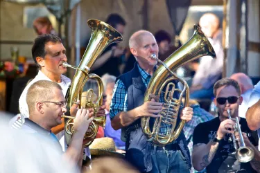 Eine Gruppe von Musikern spielt verschiedene Blechblasinstrumente auf einem Fest. Die Atmosphäre ist lebhaft und fröhlich.