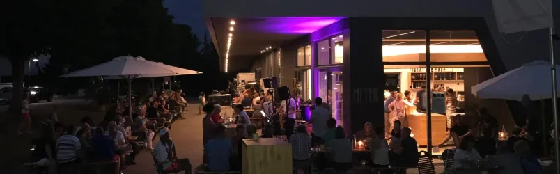 Eine belebte Bar im Freien bei Nacht, umgeben von Gästen, die entspannt sitzen. Sanftes Licht von der Bar sorgt für eine gemütliche Atmosphäre.