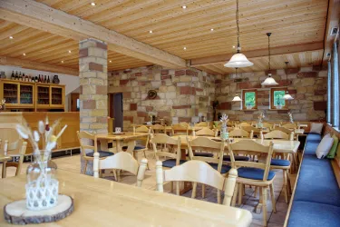 Ein gemütliches Restaurant mit Holzmöbeln und einem warmen Ambiente. Die Wände sind aus Naturstein und es gibt große Fenster für ausreichend Tageslicht.