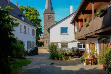 Eine malerische Gasse mit historischen Gebäuden und einer Kirche im Hintergrund. Überall sind Pflanzen und gemütliche Sitzecken zu sehen.