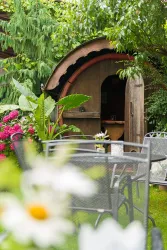 Ein gemütliches Holzhäuschen umgeben von üppigem Grün und bunten Blumen. Im Vordergrund sind schwarze Gartenmöbel sichtbar.