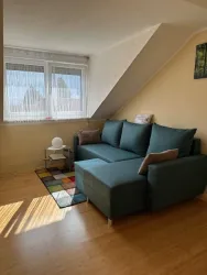 Ein gemütliches Wohnzimmer mit einem blauen Ecksofa und einem bunten Teppich. Große Fenster lassen viel Licht herein und schaffen eine freundliche Atmosphäre.