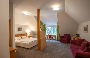 Ein Doppelbett mit Fernseher, eine rote Couch mit Sessel, ein Schreibtisch mit Stuhl und Blick in den Garten 