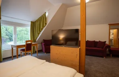 Blick vom Doppelbett auf einen Fernseher, Schreibtisch mit Stuhl vor einem Fenster und einer roten Couch