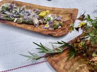 Ein Holztablett mit bunten, getrockneten Blumen und Kräutern liegt auf einem weißen Tuch. Daneben befindet sich ein weiteres Tablett mit frischen Zweigen und Blüten.