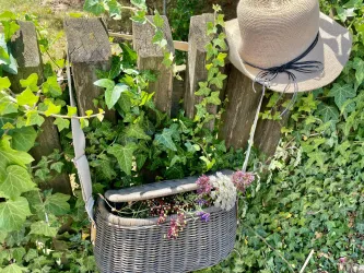 Ein Korb mit Trockenblumen hängt an einem Holzzaun, daneben liegt ein Hut. Die Umgebung ist von grünem Efeu umrahmt.