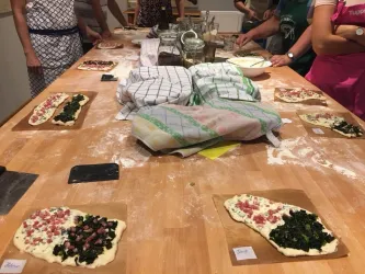Eine Arbeitsplatte voller Teigplatten mit verschiedenen Belägen. Im Hintergrund sind Menschen beim Zubereiten von Speisen zu sehen.