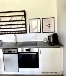 Eine moderne Küche mit einem Kühlschrank, einem Backofen und einer Kaffeemaschine. An der Wand hängen zwei Bilder und ein offenes Regal mit Geschirr.