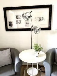 Eine moderne Stehlampe mit zwei Lichtquellen steht neben einem kleinen Tisch. Auf dem Tisch befindet sich ein Blumentopf mit einer Pflanze und im Hintergrund hängt ein gerahmtes Kunstwerk.